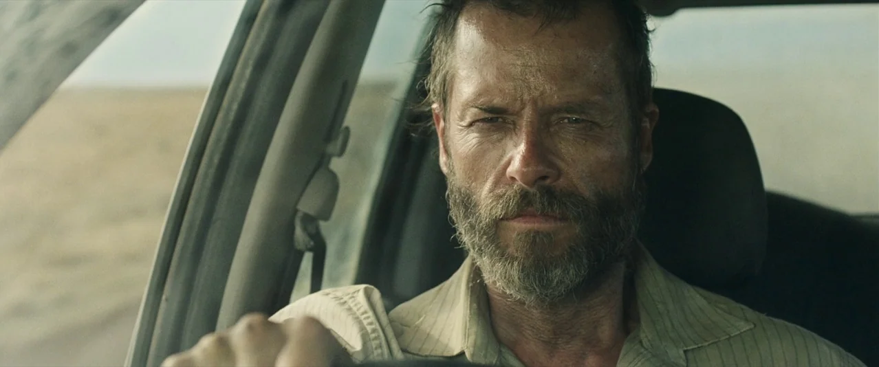 The Rover (2014) movie still: closeup — The interior of a vehicle shows a man behind the steering wheel. He looks ahead while driving through a vast…