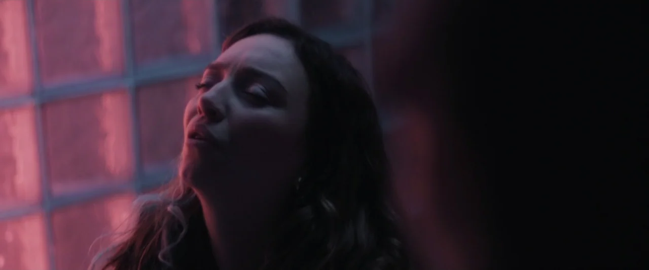 Sign Here (2017) movie still: closeup — A dark interior space features a glass block wall illuminated by bright magenta light. A young woman leans…
