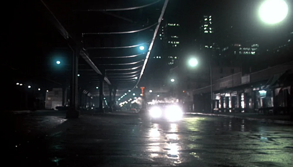 Marathon Man (1976) movie still: wide — A wet asphalt road passes under an elevated rail structure at night in a city. Bright headlights of an…