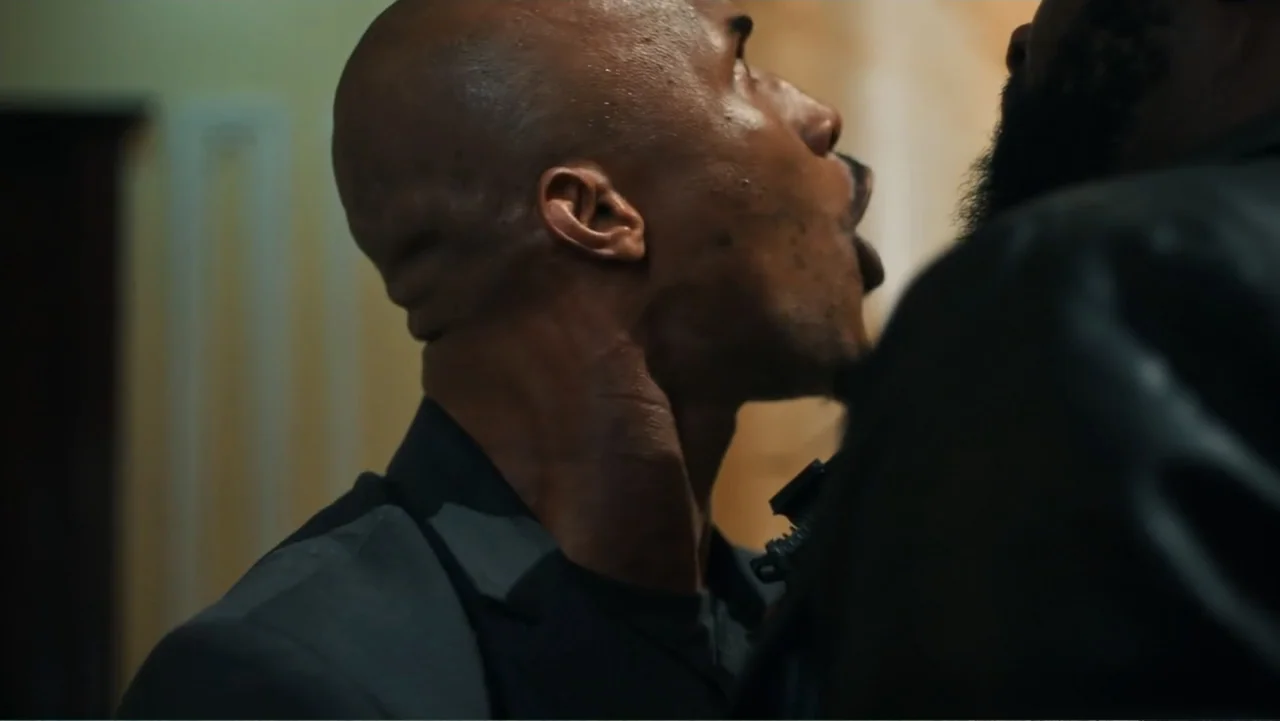 SIN: Blackout (2025) movie still: closeup — A dimly lit indoor space with neutral walls and dark shadows. A bald man faces another person in a close…