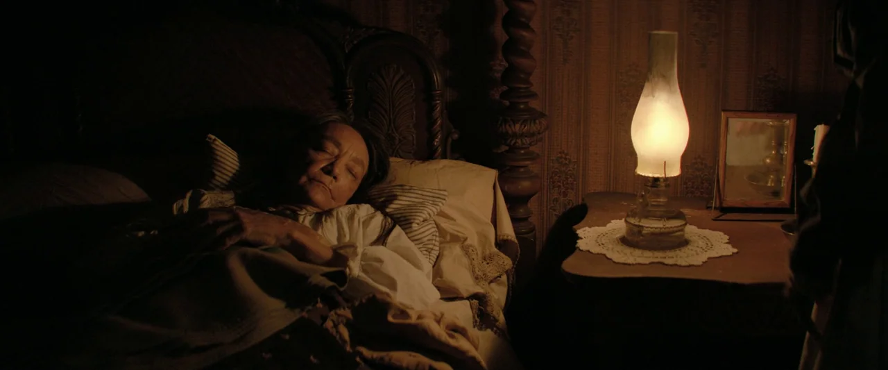 Killers of the Flower Moon (2023) movie still: medium — An aged woman lies sleeping in a four-poster bed against patterned wallpaper. A single oil…
