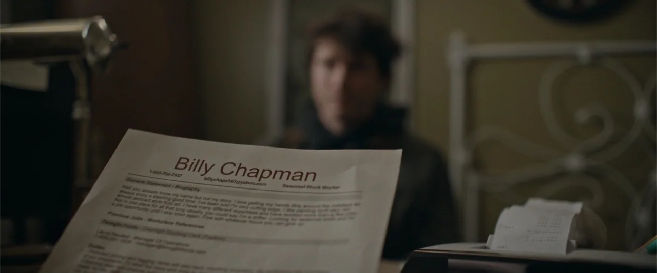 Silent Night, Deadly Night (2025) movie still: closeup — A dimly lit indoor space features a printed resume held in the foreground. The background…