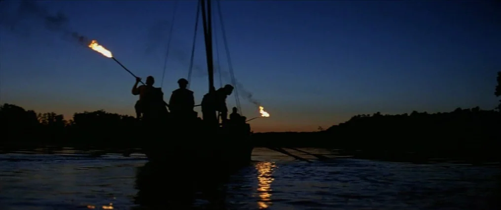 The New World (2005) movie still: wide — A dark river scene at twilight shows a wooden boat on the water. Passengers in military-style clothing hold…