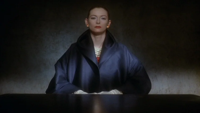 Edward II (1991) movie still: medium — A dark, dimly lit environment features a woman centered against a plain wall. The subject sits behind a dark…
