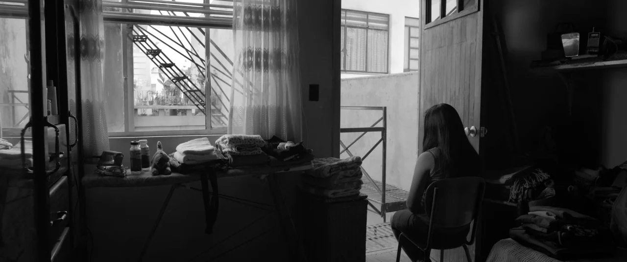 Roma (2018) movie still: wide — A cluttered room features a window overlooking an external staircase and an open door. A woman sits on a metal chair…