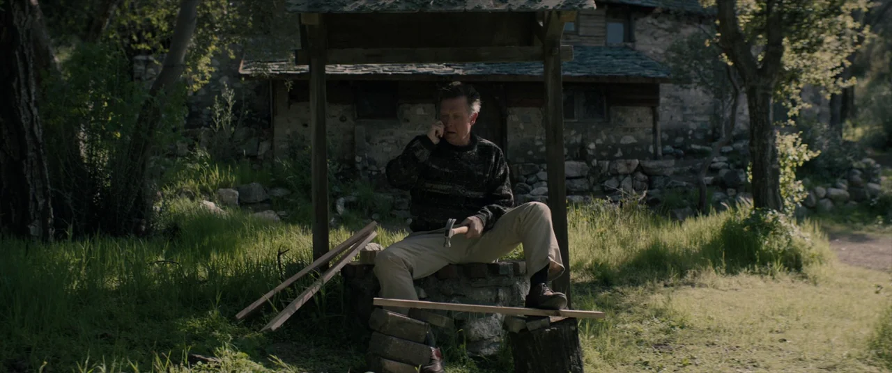 Tone Deaf (2019) movie still: fullbody — A man sits on a circular stone well structure in a grassy clearing. A small stone cottage stands in the…