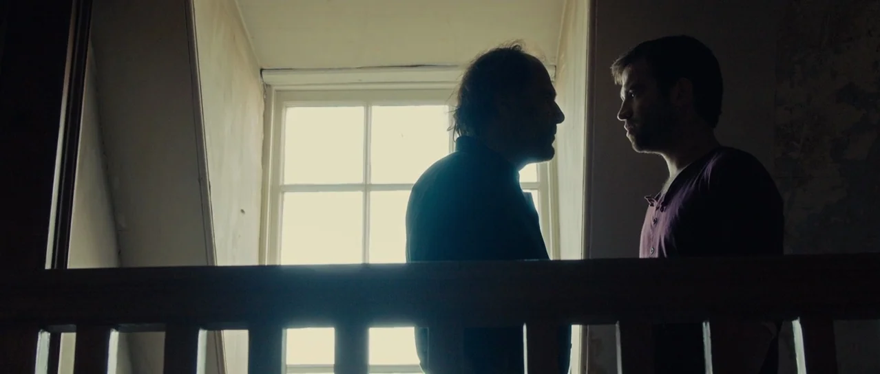 Road Games (2015) movie still: medium — A dimly lit interior landing area with a wooden banister near a bright window. Two men stand close together…