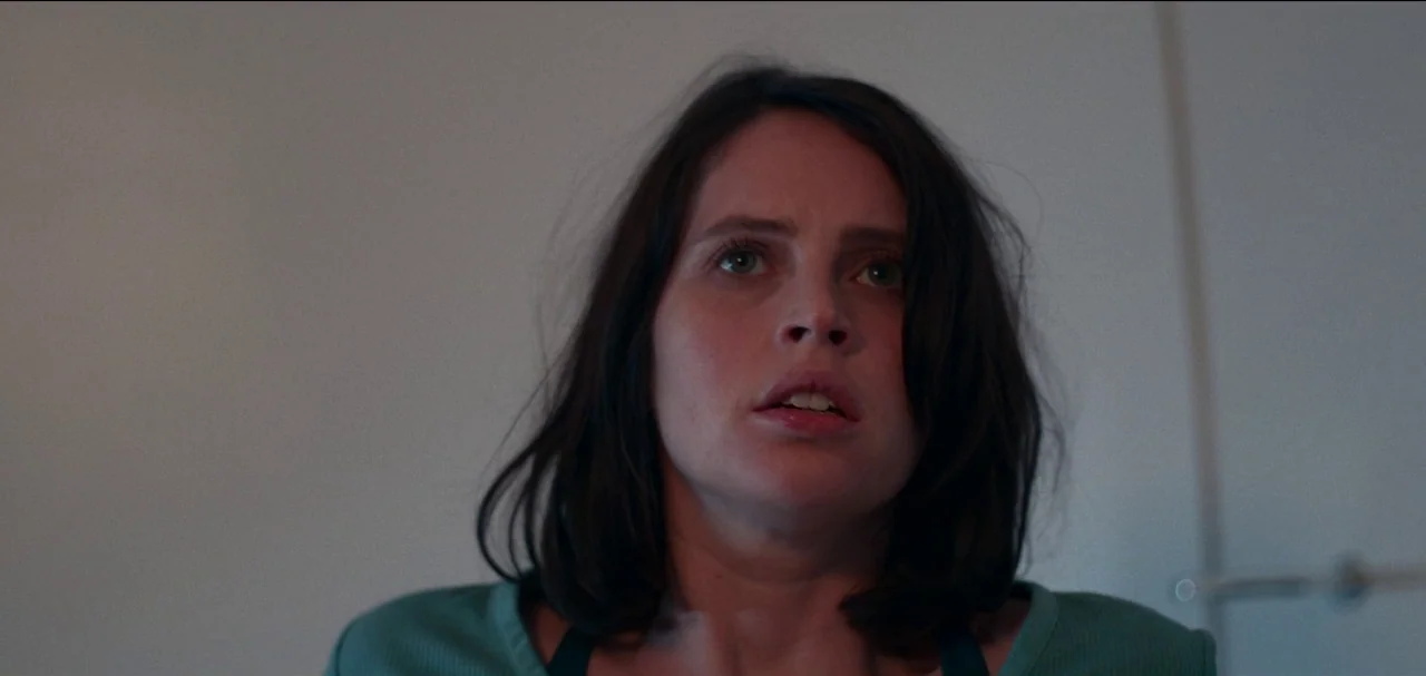 The Midnight Sky (2020) movie still: closeup — A woman stands in a plain, featureless interior space with white walls. She tilts her head slightly…