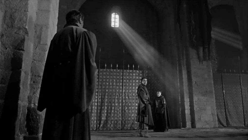 Falstaff (Chimes at Midnight) (1965) movie still: wide — A dark stone hall features tall arched ceilings and metal dividers. Two men stand in the…