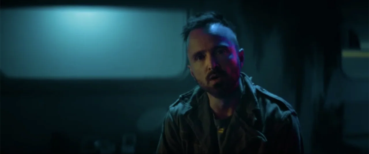 Ash (2025) movie still: medium — A dimly lit interior space with a metallic wall background and cool blue shadows. A man with a dark goatee sits in…