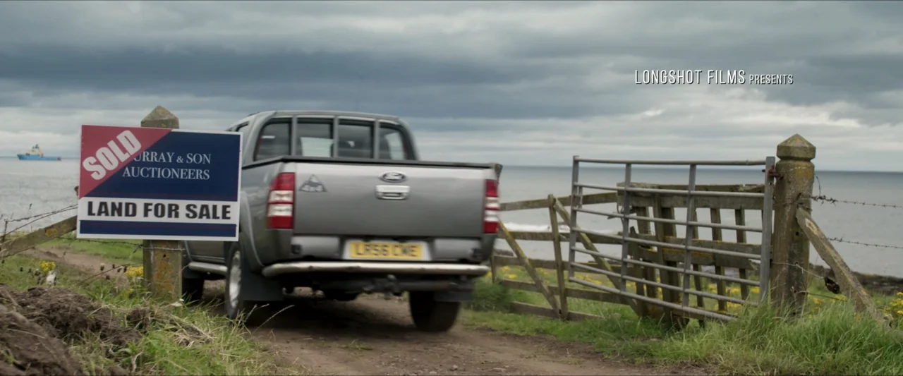 Last Breath (2025) movie still: medium — A rural coastal dirt path features a wooden gate and a sold real estate sign. A silver pickup truck travels…