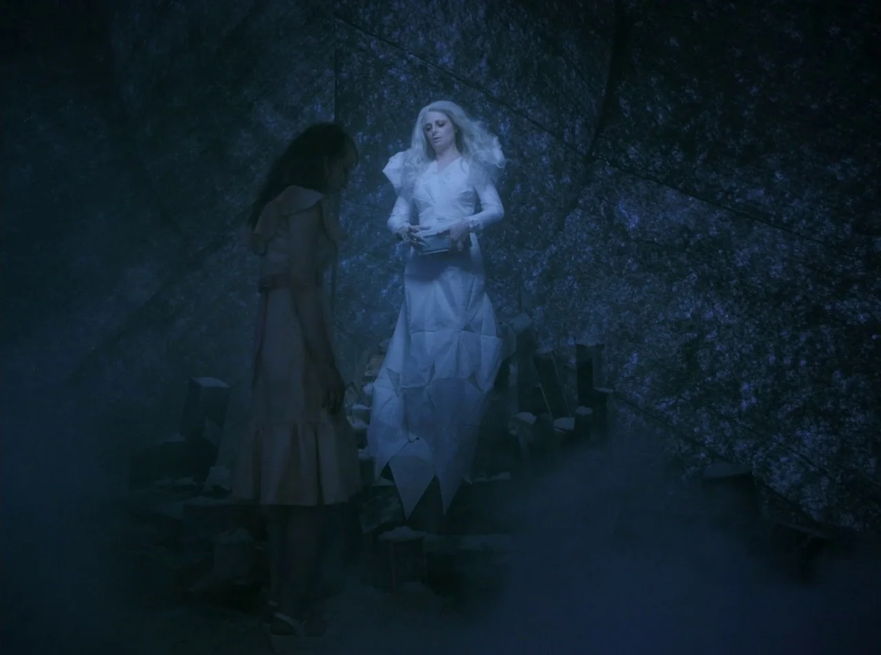 Girl Asleep (2015) movie still: fullbody — A dark, theatrical environment features low light and thick ground fog. A woman in a white gown stands…