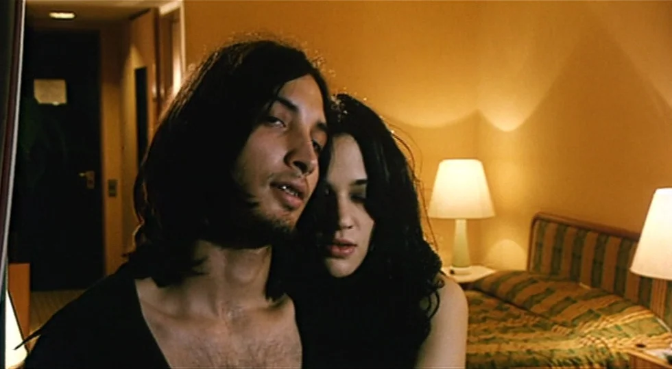 Scarlet Diva (2000) movie still: medium — A dimly lit room features a patterned bed spread and two bedside lamps. A man and a woman stand close to…