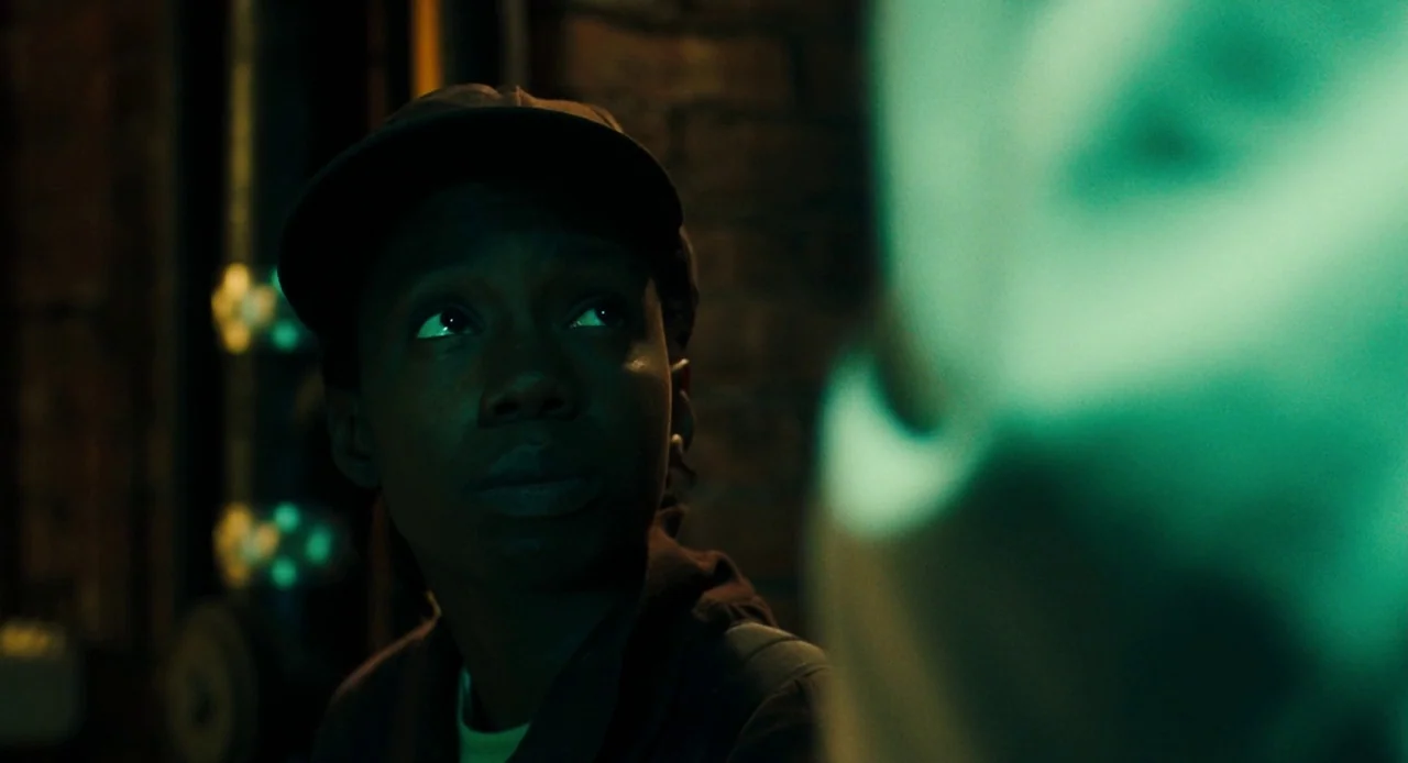 Pariah (2011) movie still: closeup — A dark, dimly lit environment features a rough brick wall and vertical pipes. A young woman in a cap looks up…