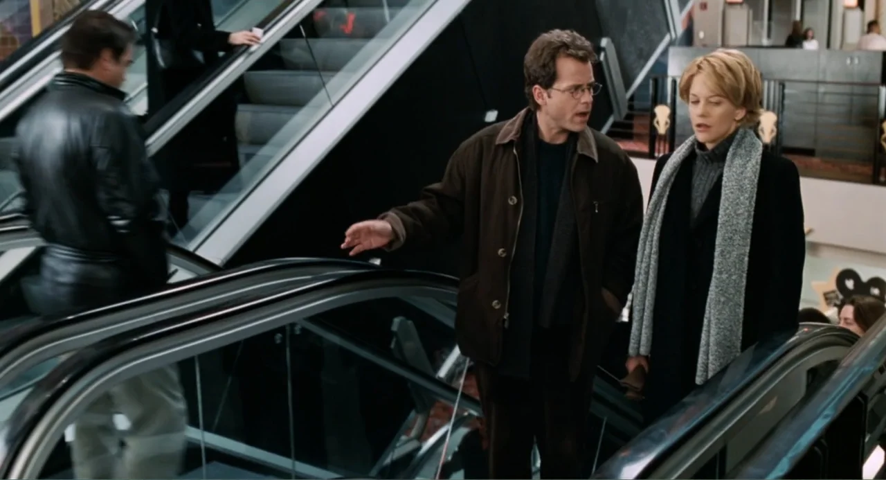 You've Got Mail (1998) movie still: medium — Two individuals stand together on an escalator inside a modern multi-level shopping center. They are…