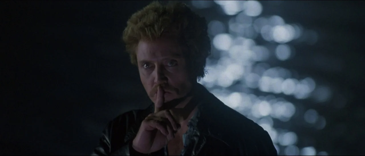 At Close Range (1986) movie still: medium — A man stands at night with out-of-focus water glinting behind him. He holds a finger to his mouth as a…