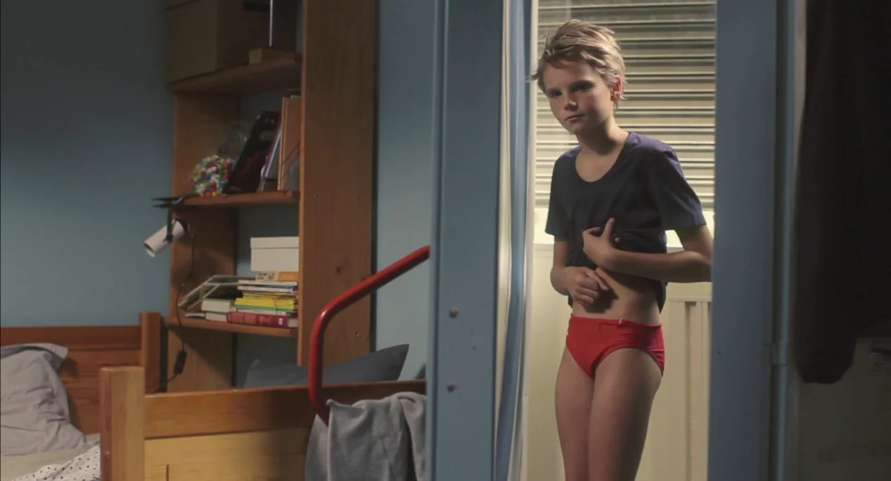 Tomboy (2011) movie still: medium — A youth stands in the doorway of a sparely furnished room with wooden shelving. The boy is examining his stomach…