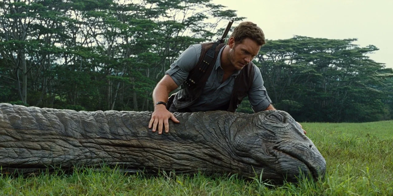 Jurassic World (2015) movie still: medium — An open grassy field with dense forest trees in the background under overcast skies. A man in a brown…