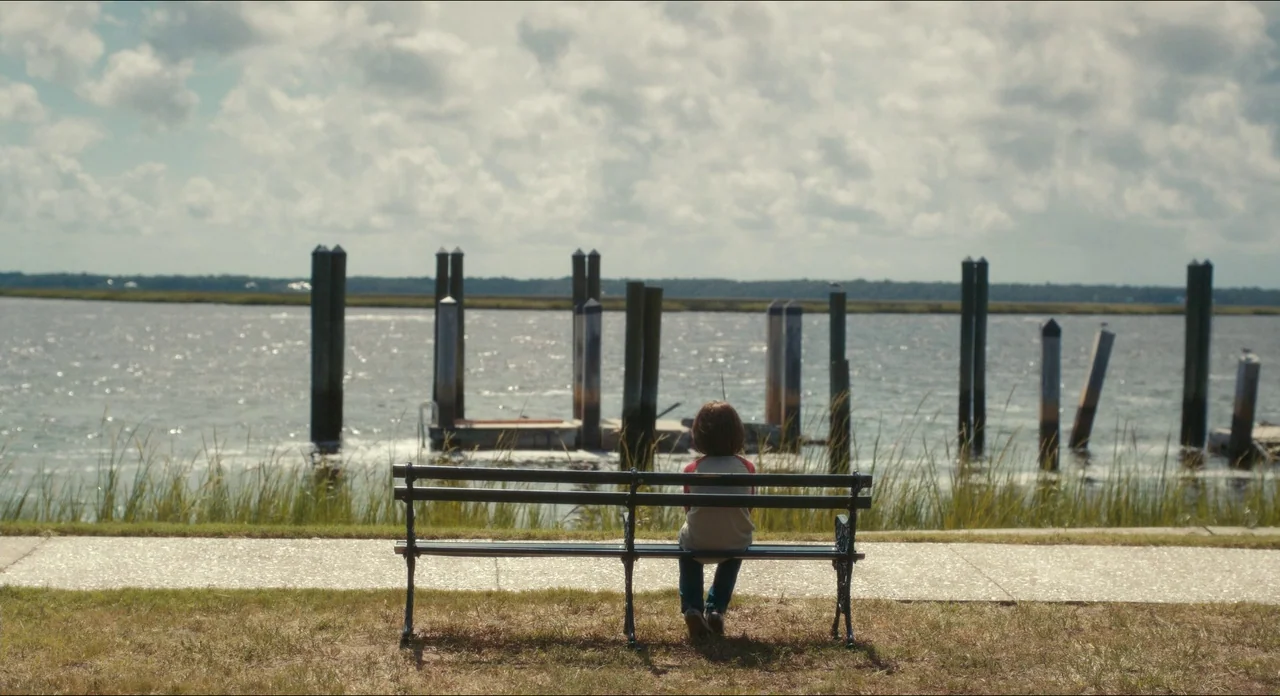 Doctor Sleep movie still: wide — A public park area overlooks a wide river with wooden pilings in the water. A young child sits alone on a dark…