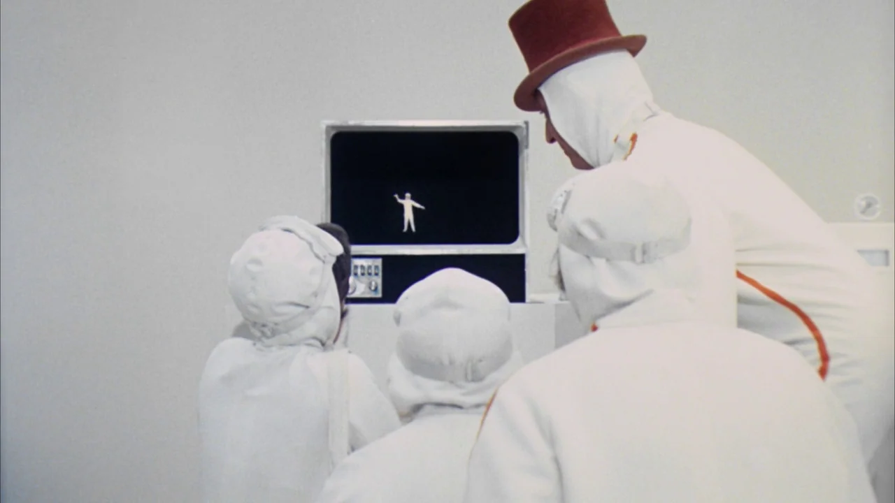 Willy Wonka & the Chocolate Factory (1971) movie still: medium — A stark white interior with a central television screen mounted on a pedestal. Four…