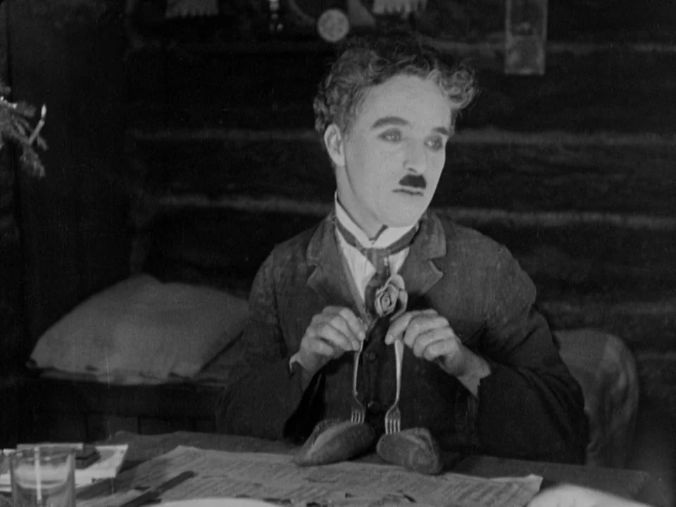 The Gold Rush (1925) movie still: medium — A rustic interior with dark log walls provides the setting for a solitary meal. The man balances two…