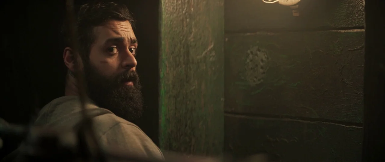 Sea Fever (2019) movie still: medium — A man stands in a dimly lit, narrow space with dark, textured green wooden walls. He looks back over his…