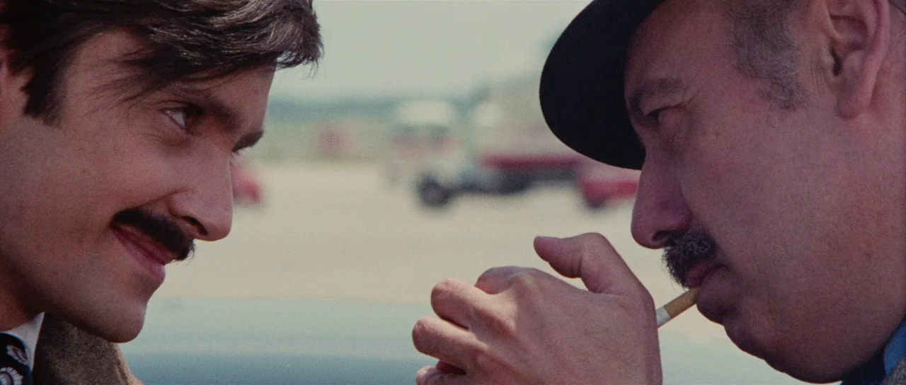 Death Walks On High Heels (1971) movie still: closeup — A sunny outdoor tarmac area provides the backdrop for two men facing each other. The younger…