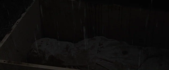 To Dust - Nightmares (2018) movie still: medium — A rain-slicked wooden crate sits in a dark, muddy pit at night. A shrouded body lies motionless…