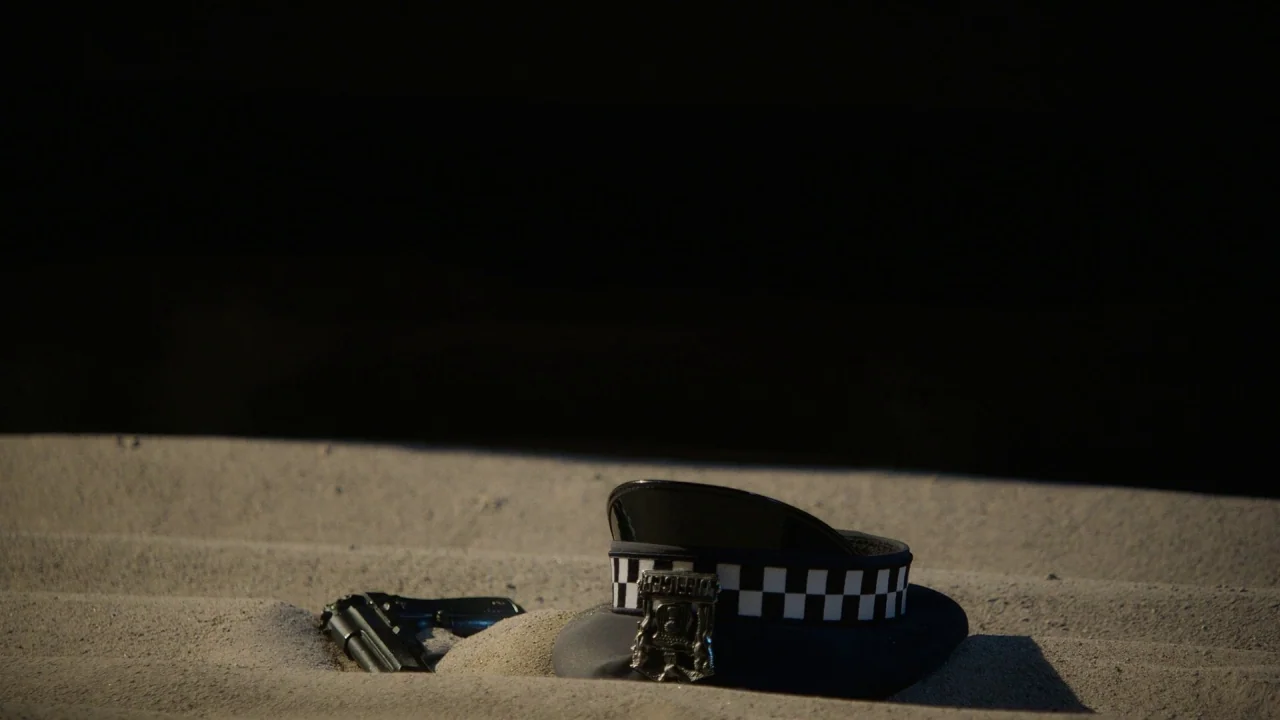 Pass Over (2018) movie still: closeup — A police cap and a semi-automatic handgun are partially buried in light-colored sand. The background is…