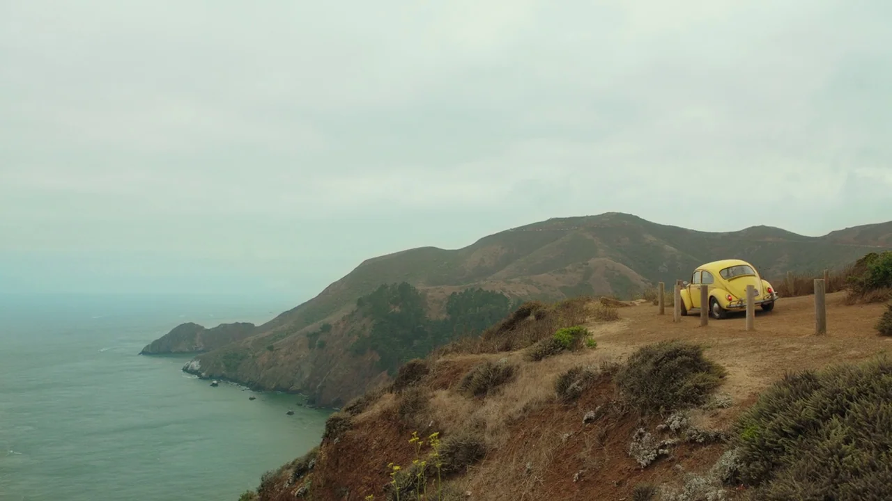 Bumblebee (2018) movie still: wide — A yellow car is parked on a dirt shoulder at the edge of a cliff. The Pacific Ocean sits below the cliff with…