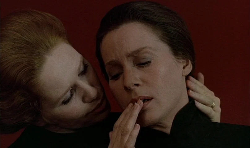 Cries and Whispers (1972) movie still: closeup — Two women stand closely against a solid dark red wall background. One woman gently touches the…