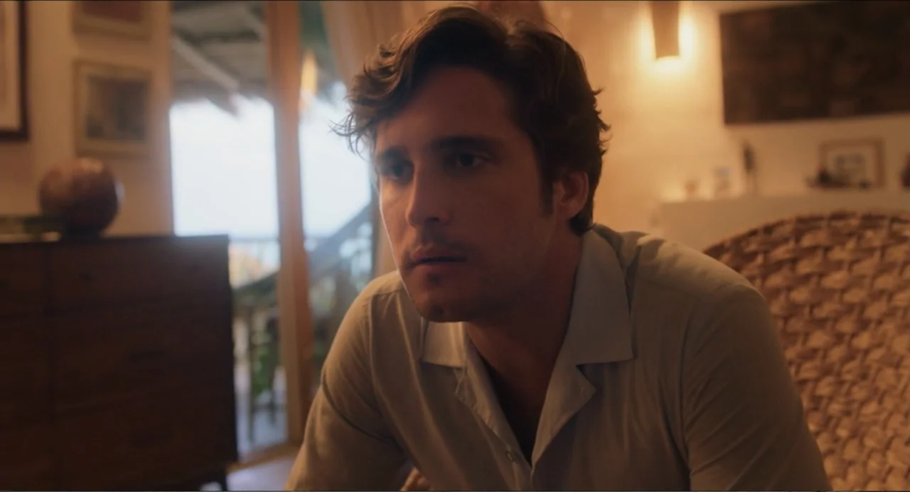 At Midnight (2023) movie still: medium — A residential room features a dark wood dresser and a woven wicker chair. A young man sits in the chair,…
