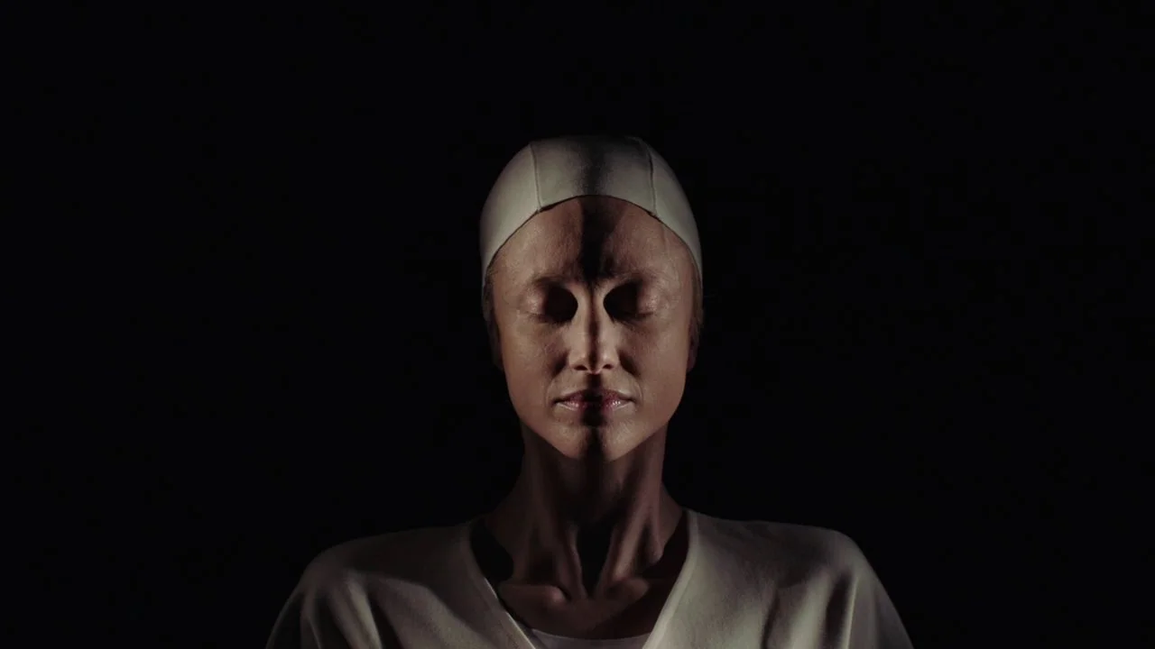 Possessor (2020) movie still: closeup — A featureless black background surrounds a single person centered in the frame. The subject wears a plain…
