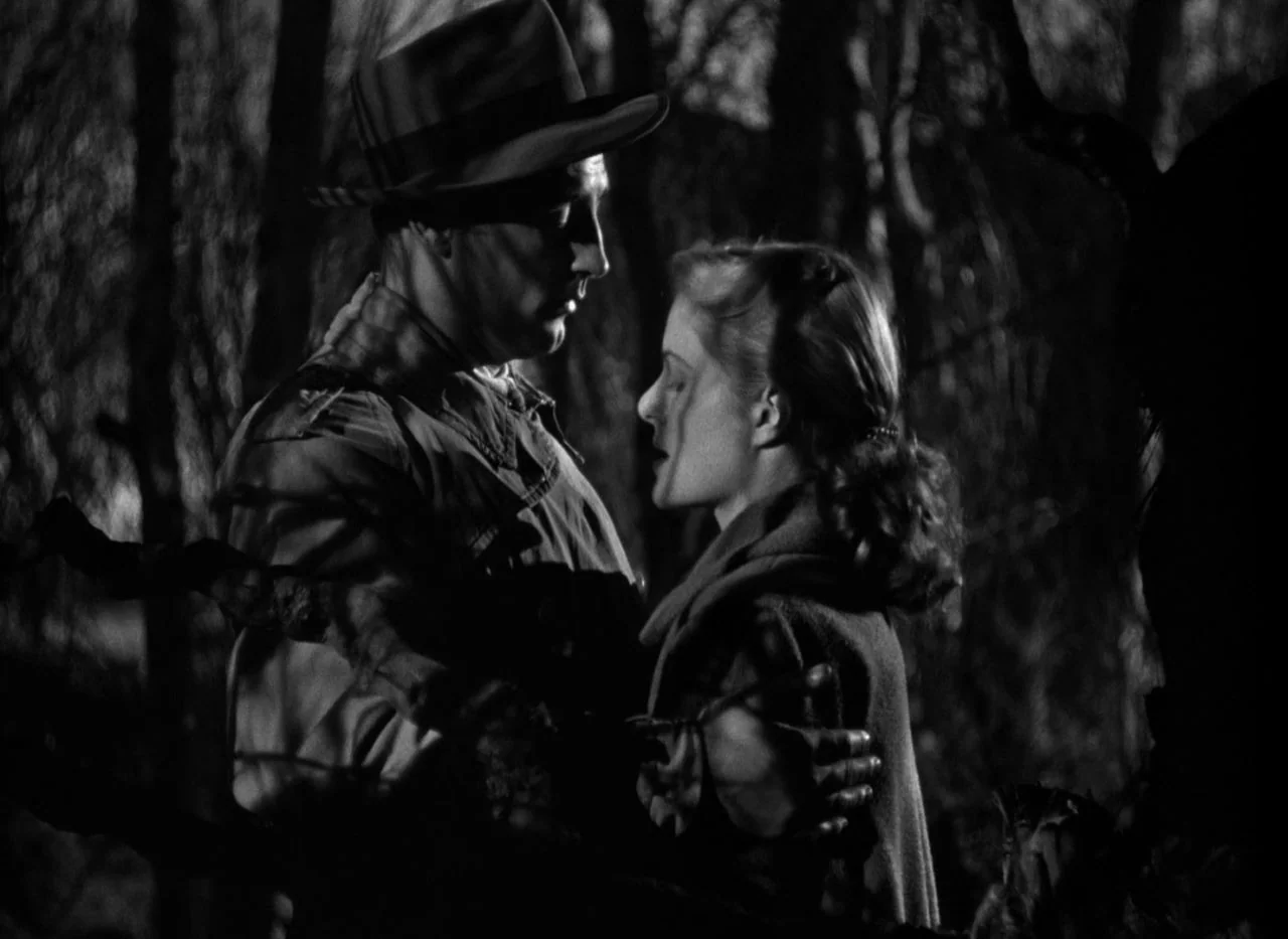 Out of the Past (1947) movie still: medium — A man and a woman stand together in a dark, densely wooded area at night. The pair are positioned…