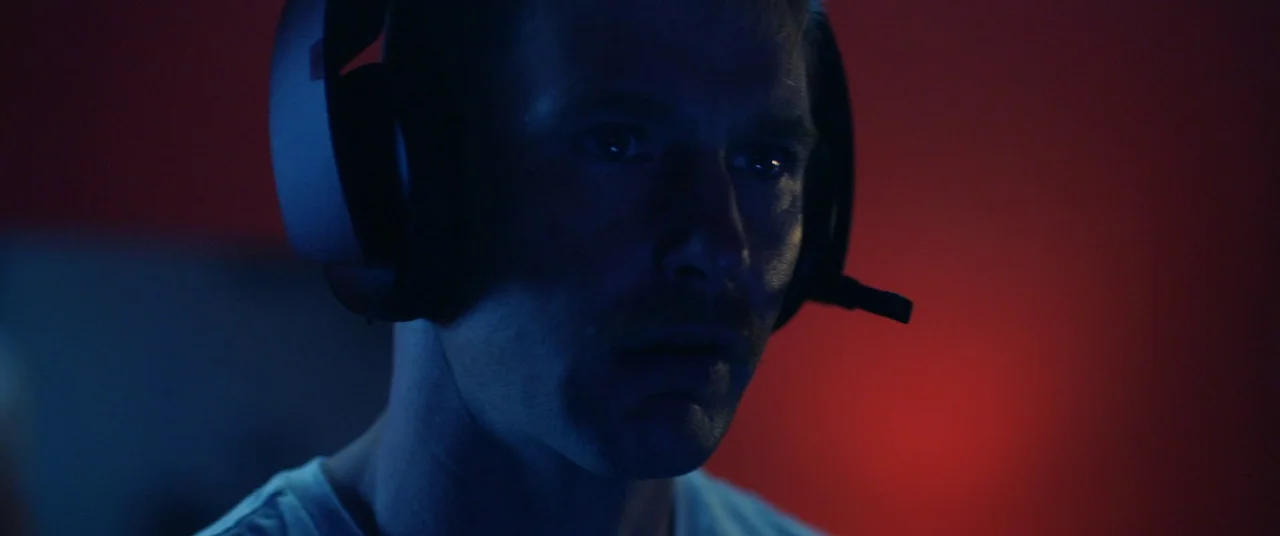 Blindfire (2020) movie still: closeup — A man wearing headset hardware sits in a dark room illuminated by electronic light. He stares forward while…