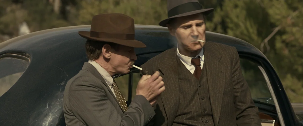 Marlowe (2023) movie still: medium — Two men in suits and fedoras stand before a dark vintage sedan outdoors. One man uses a lighter to ignite a…