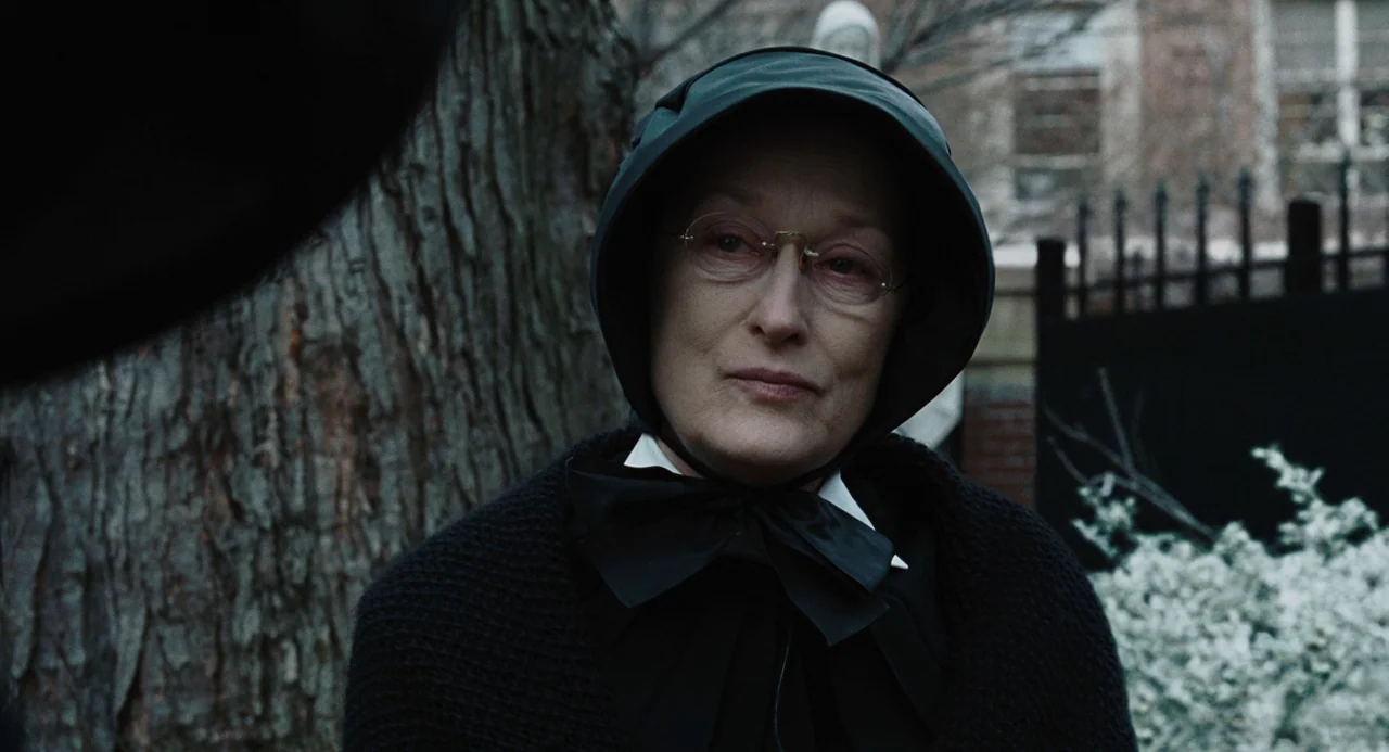 Doubt (2008) movie still: closeup — A woman stands in a muted outdoor courtyard with a tree and iron fence. She looks toward an unseen individual…