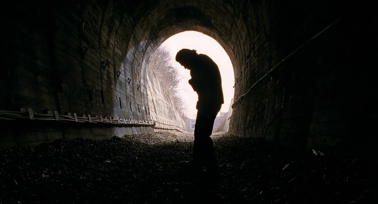 I Saw The Devil (2010) movie still: fullbody — A concrete tunnel with a gravel floor leads to an illuminated outdoor opening. A man stands in…