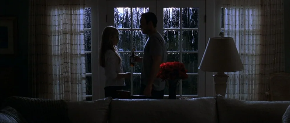 American Beauty (1999) movie still: wide — An interior space with a glass patio door showing rain against the dark exterior glass. A couple stands…