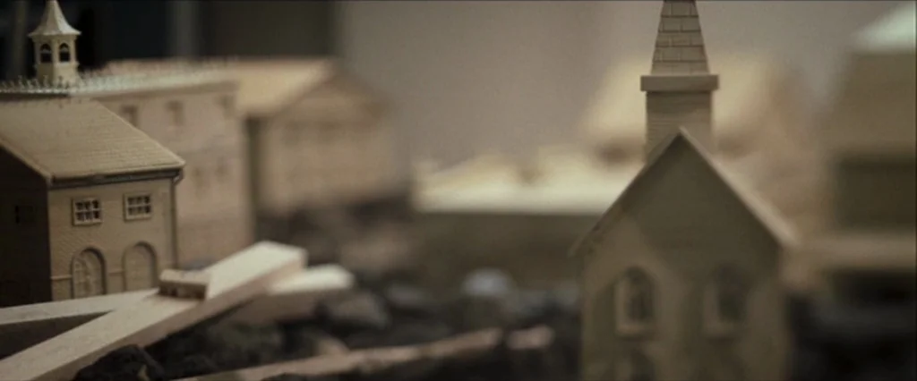 Moon (2009) movie still: closeup — A miniature town model features small wooden buildings arranged on a dark gravel base. The structures include a…