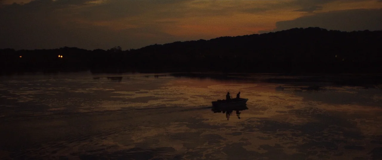 Dark Waters (2019) movie still: wide — A wide shot shows a calm, dark lake at dusk with rolling forested hills. Two figures operate a small boat,…