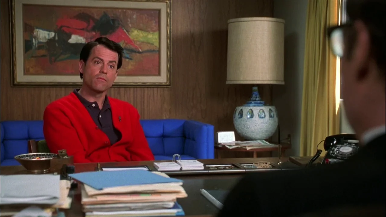 Auto Focus movie still: medium — A 1960s wood-paneled room features blue furniture, an abstract painting, and a desk lamp. A man in a bright red…