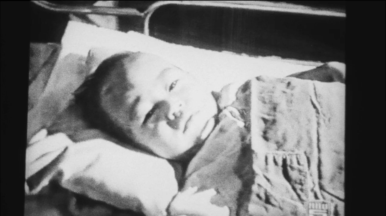 Mysterious Object at Noon (2000) movie still: closeup — An infant rests on a pillow inside a metal crib structure. The baby lies still and gazes…