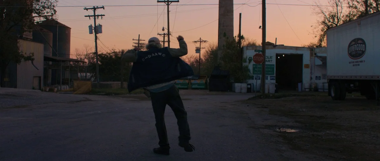Joe (2013) movie still: fullbody — An asphalt road runs between industrial storage buildings, silos, and a large shipping truck. A man stands in the…