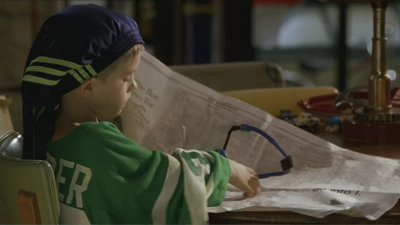 Big Daddy (1999) movie still: medium — A child sits at a wooden table inside a dimly lit interior space. The boy looks down at a large spread…