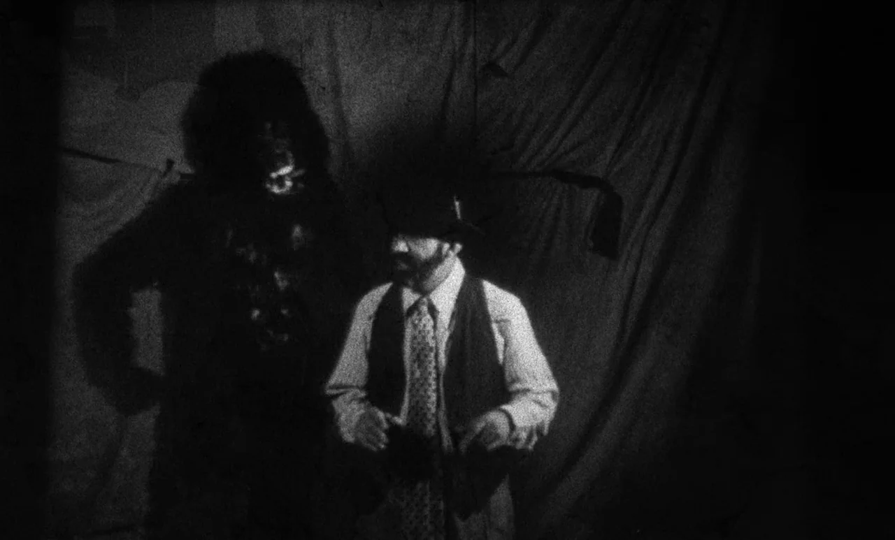 Go Down Death movie still: medium — A dark, makeshift film set features heavy curtains in the background. A man in a suit stands next to a person…