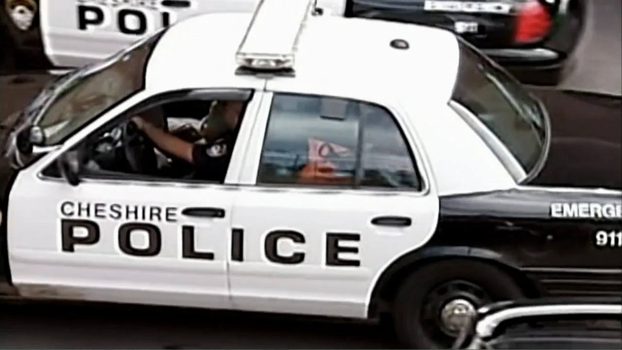 The Cheshire Murders (2013) movie still: medium — A marked police sedan travels along a paved asphalt road during daylight hours. A police officer…