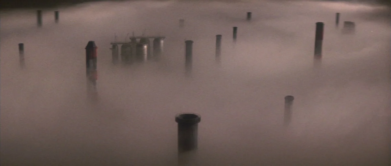 Godzilla Vs Hedorah (1971) movie still: establishing — An industrial area with numerous vertical pipes is obscured by a dense layer of fog. The…