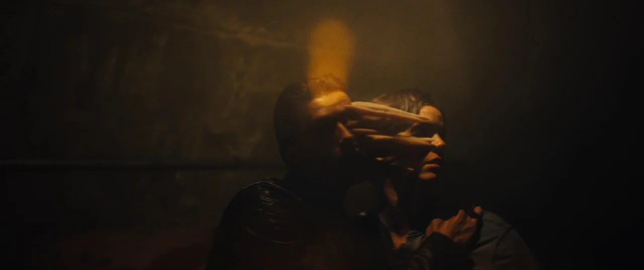 Daniel Isn’t Real movie still: medium — A dark, indistinct interior space is filled with soft golden light. One person holds their hand firmly over…