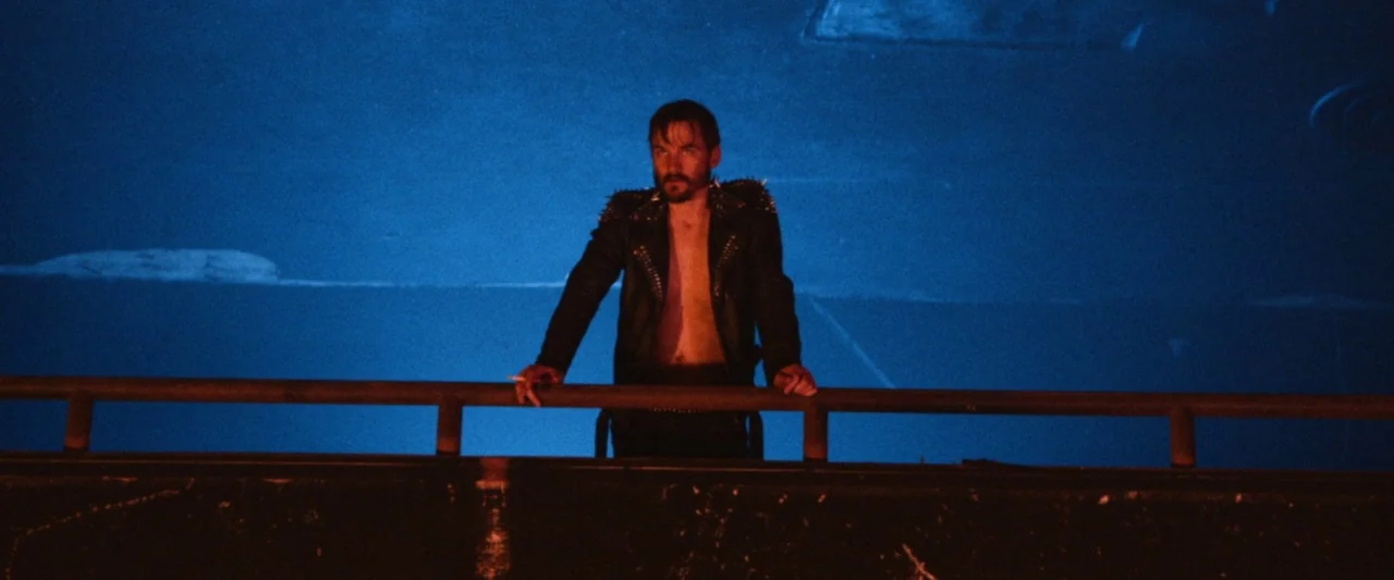 VFW movie still: medium — A man stands behind a dark metal railing above a brightly lit blue surface. The man is wearing an unzipped leather jacket…